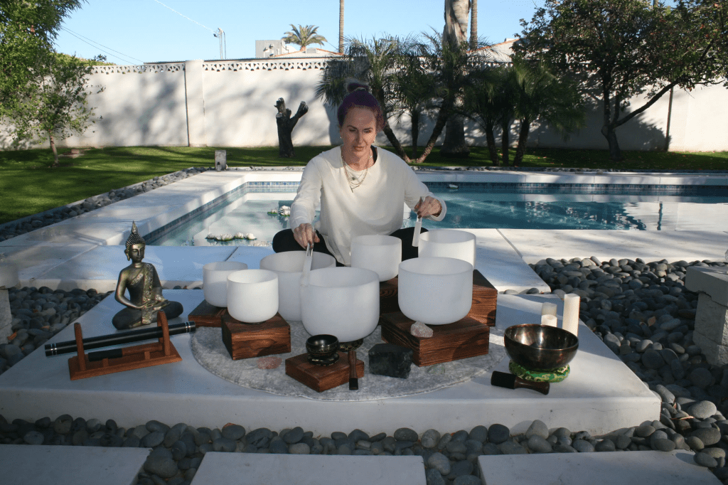 A woman with purple hair playing crystal singing bowls in a tranquil garden setting near a swimming pool, accompanied by a small Buddha statue and various musical instruments.